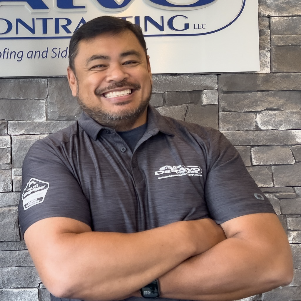 Smiling contractor in gray polo shirt with crossed arms standing in front of stone wall and contracting company sign