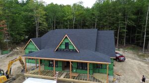 House under construction with dark shingles, green weatherproofing, wooden frame porch, and two triangular dormers in wooded setting