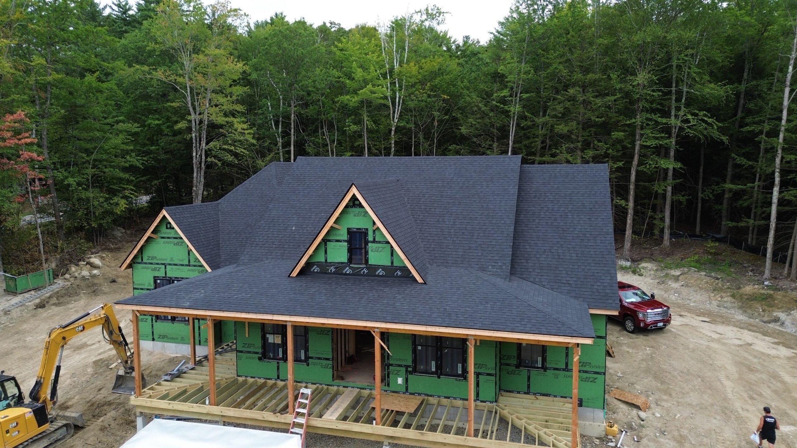 House under construction with dark shingles, green weatherproofing, wooden frame porch, and two triangular dormers in wooded setting