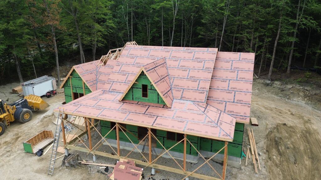 Aerial view of house under construction with pink insulation sheathing on roof, green wall sheathing, wooden frame, and construction equipment