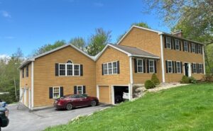 Two-story colonial house with tan vinyl siding, black shutters, and garage. Red car parked in driveway on green lawn with trees.