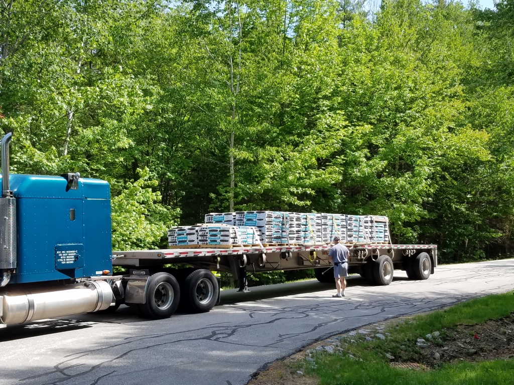 Blue semi-truck with flatbed trailer loaded with stacked construction materials parked on residential street with green trees