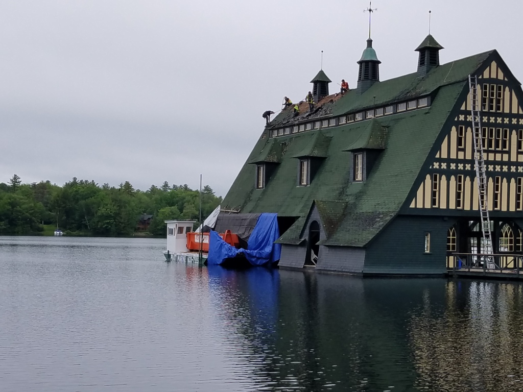 Historic Tudor-style boathouse with green roof and cupolas sits on lake with boats covered in blue tarps, workers on roof