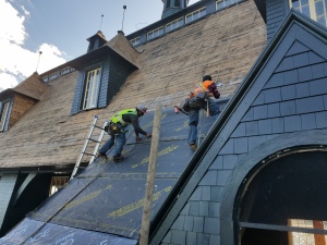 Two roofers in safety gear installing blue underlayment on steep residential roof with ladder, working on home construction project
