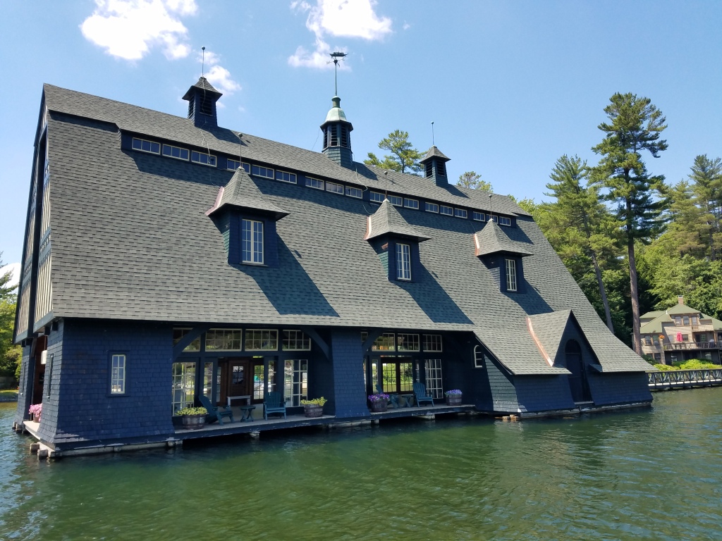 Large blue boathouse with gray shingled roof and multiple dormers floating on lake water, surrounded by green trees under blue sky