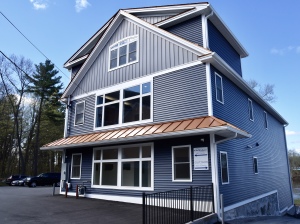 Modern two-story house with gray siding, white trim, copper roof overhang, and large windows surrounded by trees