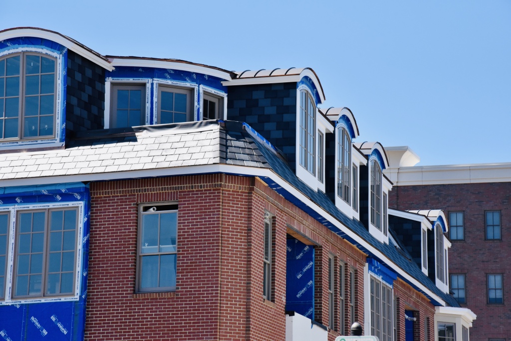 New construction townhouses with red brick facades and blue house wrap, featuring white-framed windows and dormers under clear blue sky