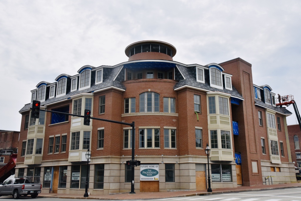 Multi-story brick building with curved corner design, dormers, and glass cupola under construction in downtown area