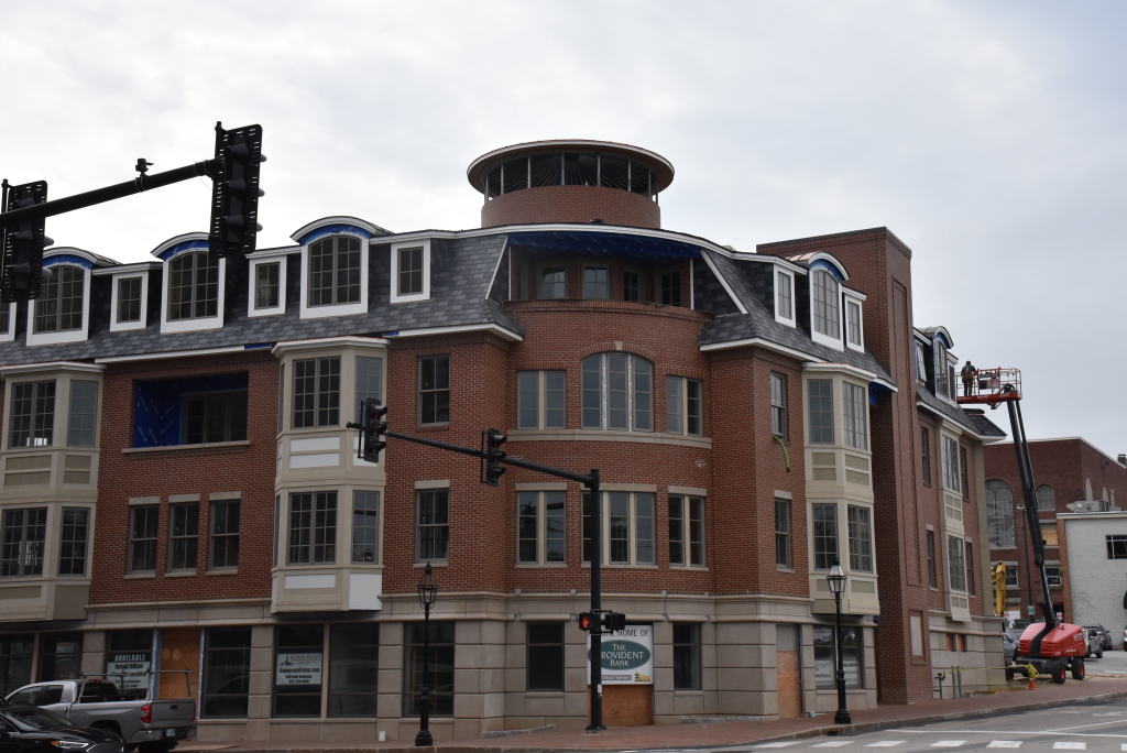 Modern brick building with curved corner facade, featuring mansard roof with dormers and ground-floor retail spaces on urban street corner