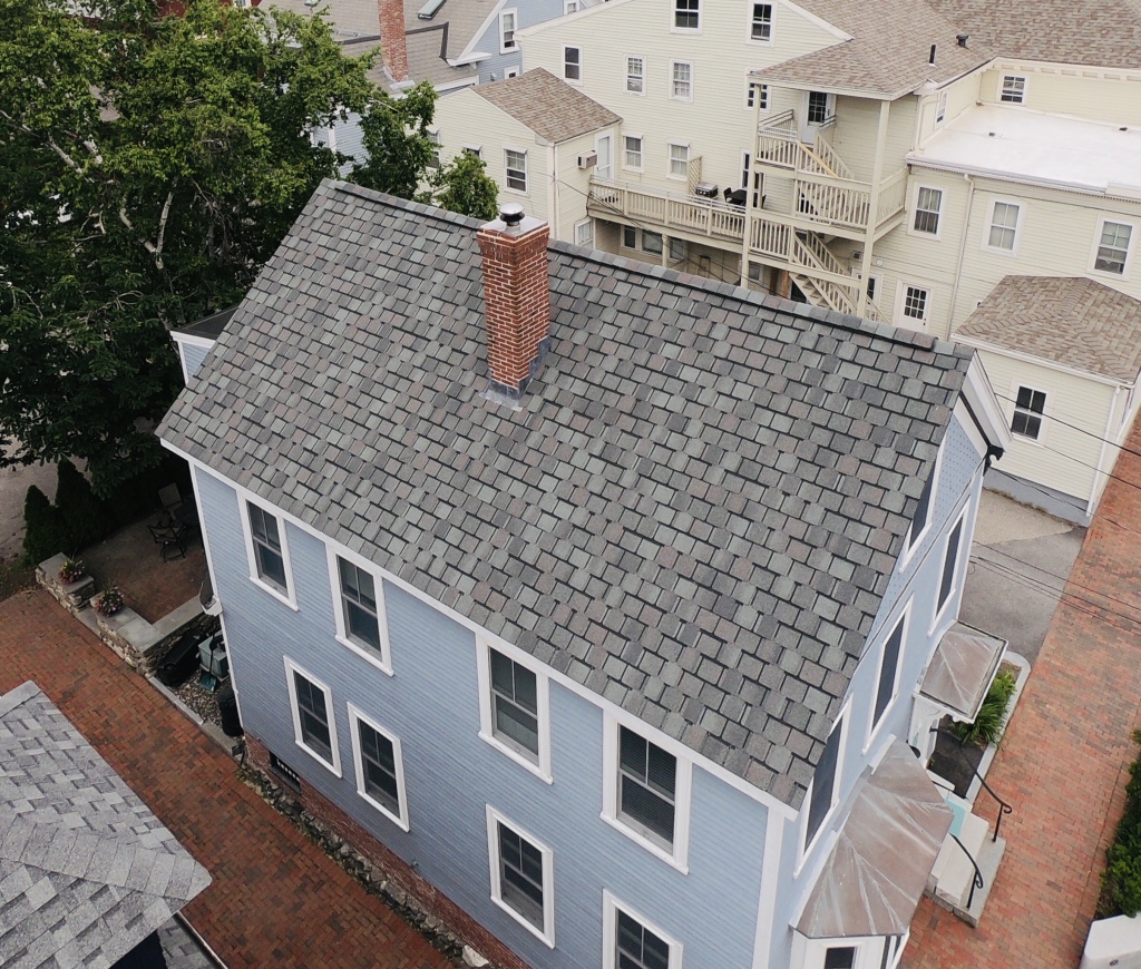 Aerial view of blue colonial house with gray asphalt shingle roof and red brick chimney in historic neighborhood