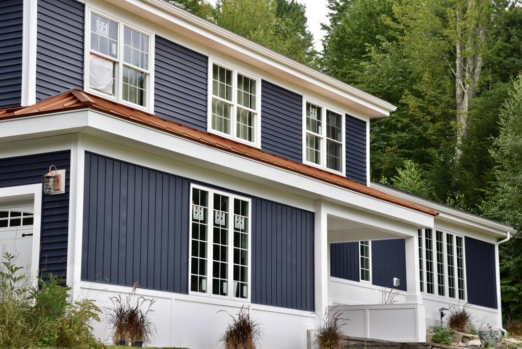 Two-story house with navy blue siding, white trim, copper roof accents, and large windows surrounded by lush green trees