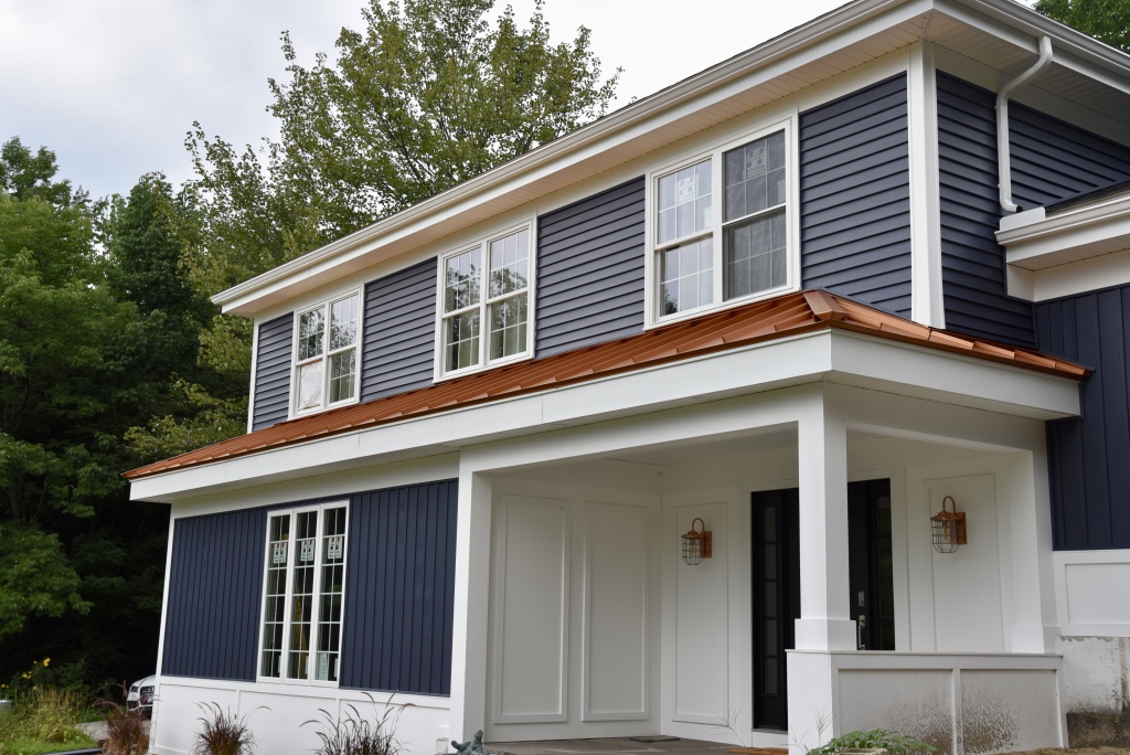 Modern two-story house with navy blue and white siding, copper awning, white columns, and front porch surrounded by trees