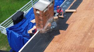 Aerial view of roofers installing black underlayment and wood shingles around brick chimney on residential roof construction project