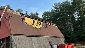 Construction workers on steep roof installing yellow underlayment and shingles on residential building with forest background