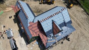 Aerial view of house under construction showing exposed roof trusses, blue underlayment, brick walls, and construction equipment on dirt lot