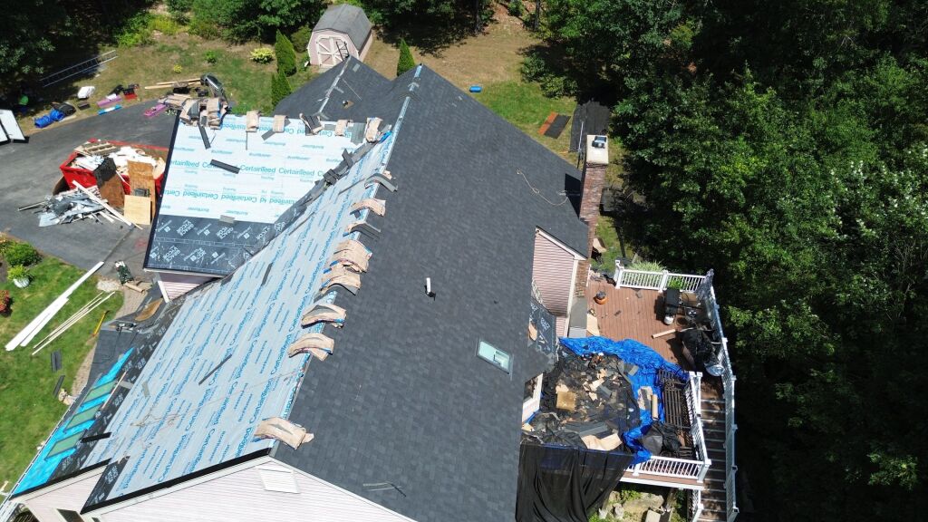 Aerial view of house roof replacement in progress showing new dark shingles, blue underlayment, and construction materials scattered around