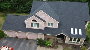 Aerial view of pink house with dark gray shingles, dormer windows, three-car garage, and landscaped yard surrounded by trees