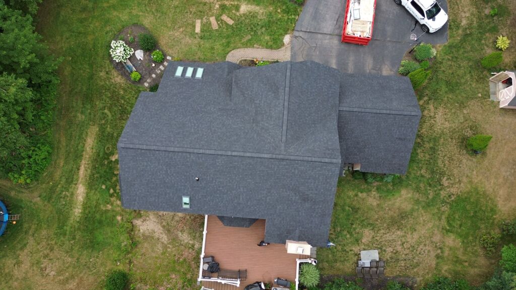 Aerial view of residential home with dark gray shingle roof, wooden deck, landscaped yard, and driveway with white car