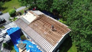 Aerial view of house under construction with exposed wooden roof rafters, blue tarps covering materials, surrounded by green trees