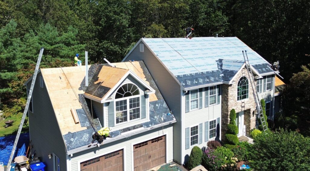 Aerial view of house undergoing roof replacement with workers installing new shingles over protective underlayment on residential home