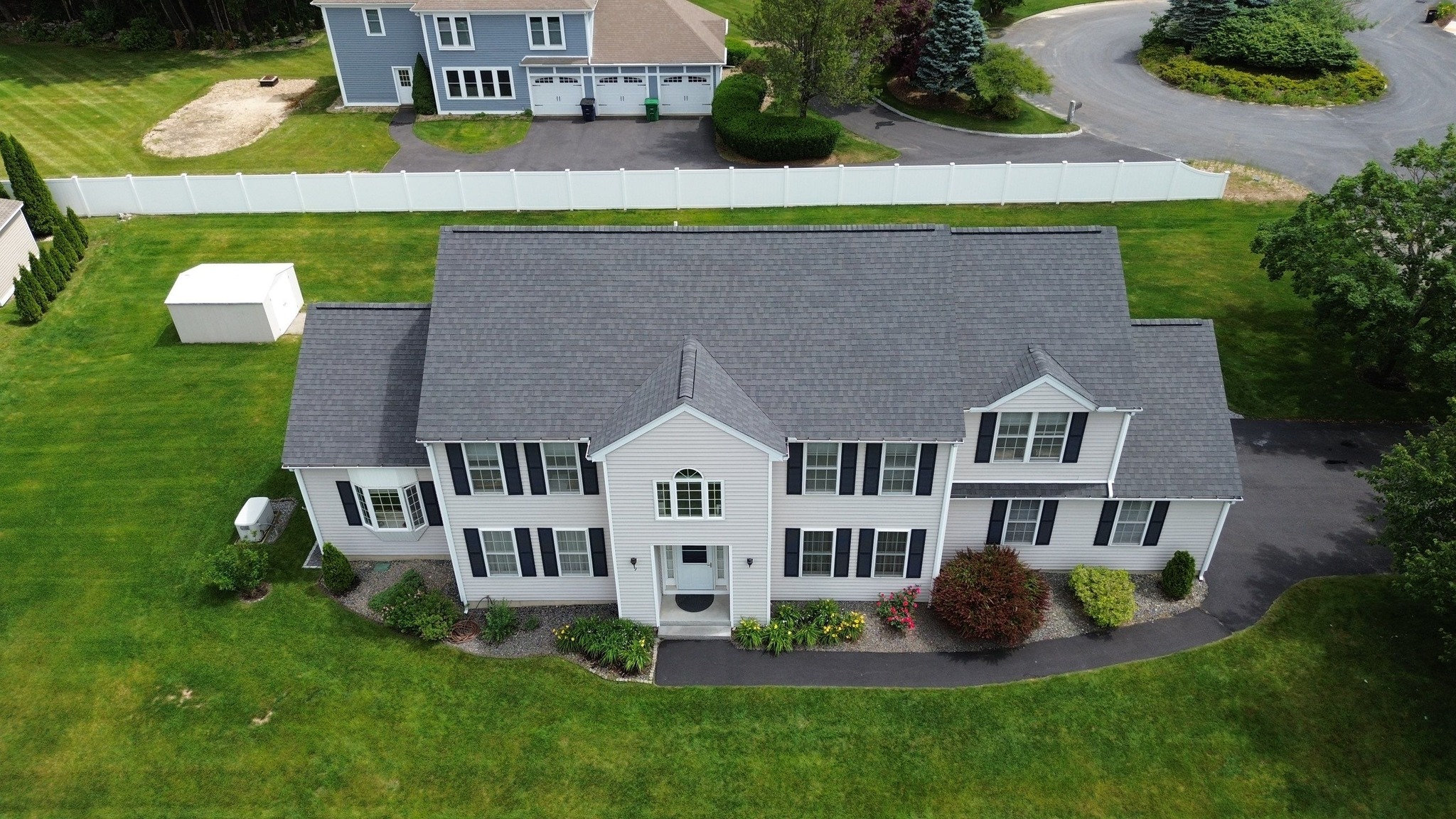 Aerial view of white colonial house with dark shutters, gray roof, circular driveway, and landscaped yard in residential neighborhood