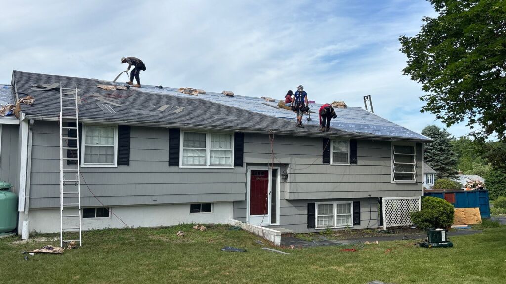 Roofing contractors installing new shingles on gray split-level house with ladder against side and materials scattered on roof