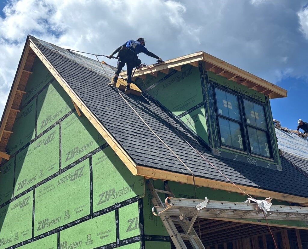 Construction worker installing roof shingles on house with green ZIP System sheathing and dormer windows under cloudy sky