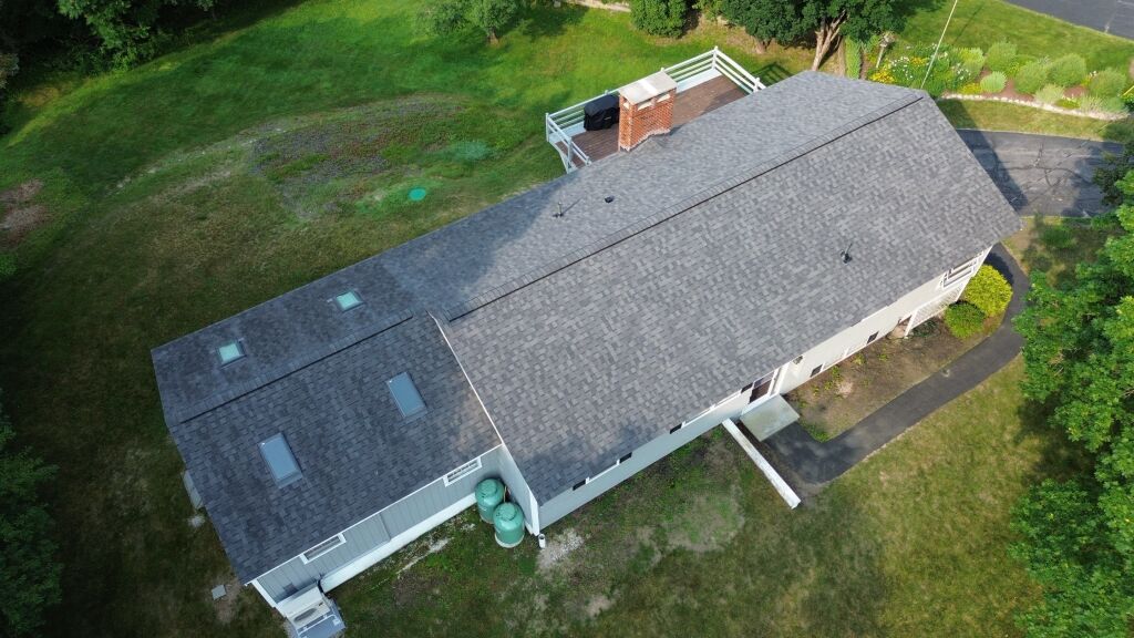 Aerial view of single-story house with dark gray shingle roof, skylights, brick chimney, and white gutters surrounded by green lawn
