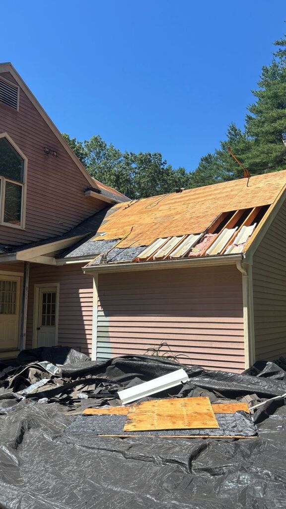 House roof under construction with exposed wooden sheathing, torn shingles, and tarps on ground during roofing renovation project