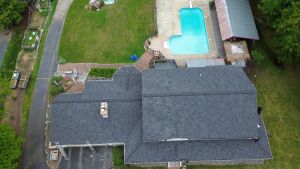 Aerial view of residential property with dark gray shingle roof, backyard swimming pool, and surrounding green lawn and trees