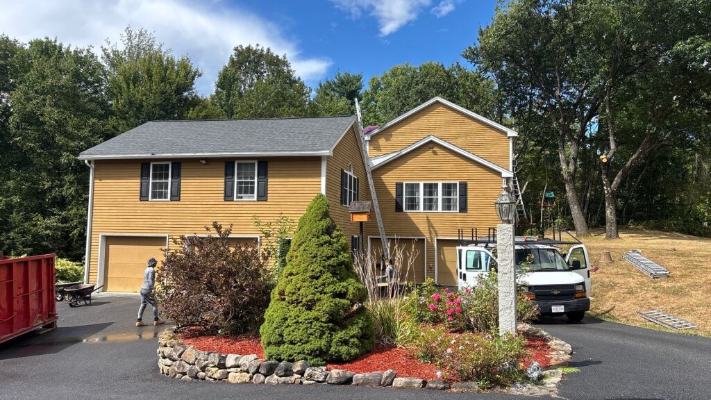 Yellow vinyl siding house with garage, landscaped garden, utility trucks in driveway, surrounded by trees on sunny day