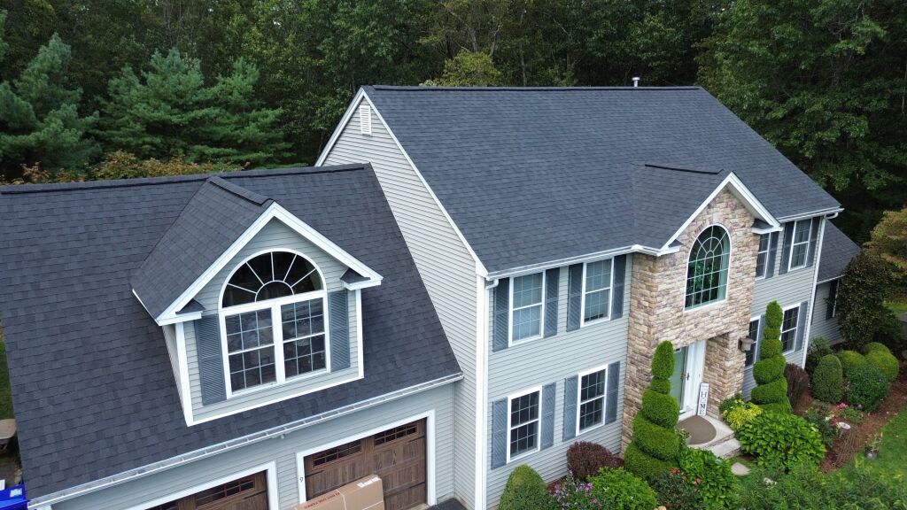 Aerial view of large two-story house with gray shingle roof, beige vinyl siding, stone accents, attached garage, and landscaped front yard
