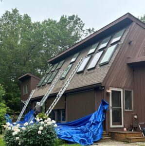 Workers installing skylights on brown wooden house roof using extension ladders, with blue tarps protecting landscaping below