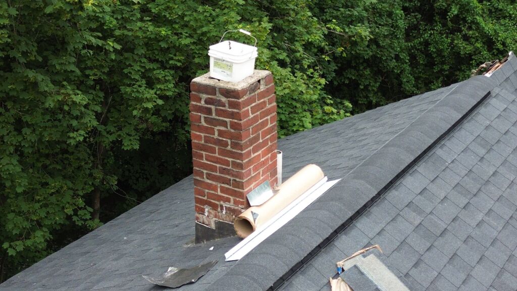 Brick chimney on gray shingled roof with white chimney cap and copper flashing, surrounded by green trees