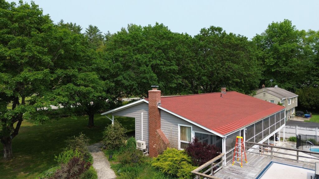 Aerial view of single-story house with red metal roof, brick chimney, and screened porch surrounded by lush green trees