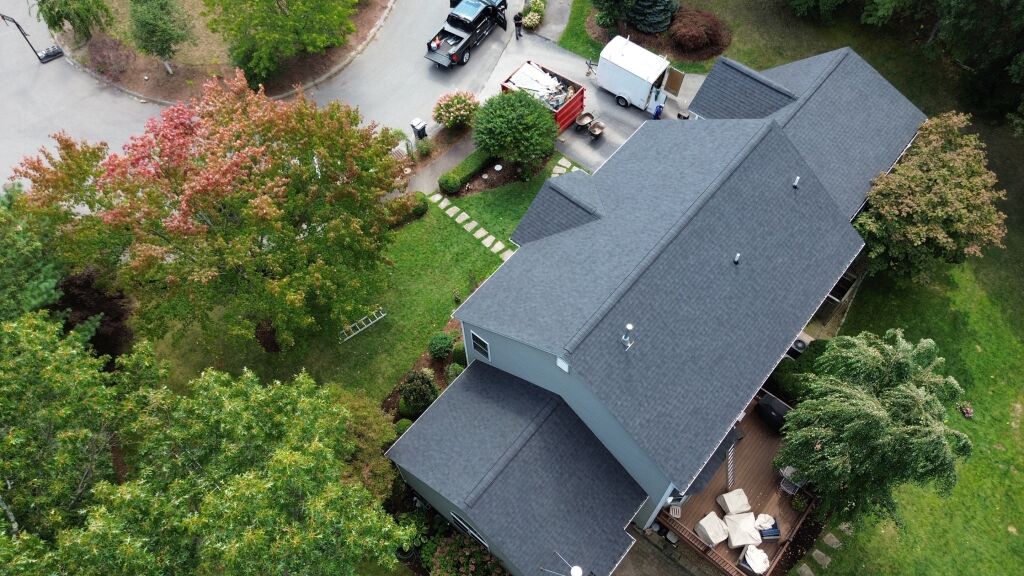 Aerial view of house with dark gray shingle roof, landscaped yard with colorful trees, stone walkway, and moving trucks in driveway