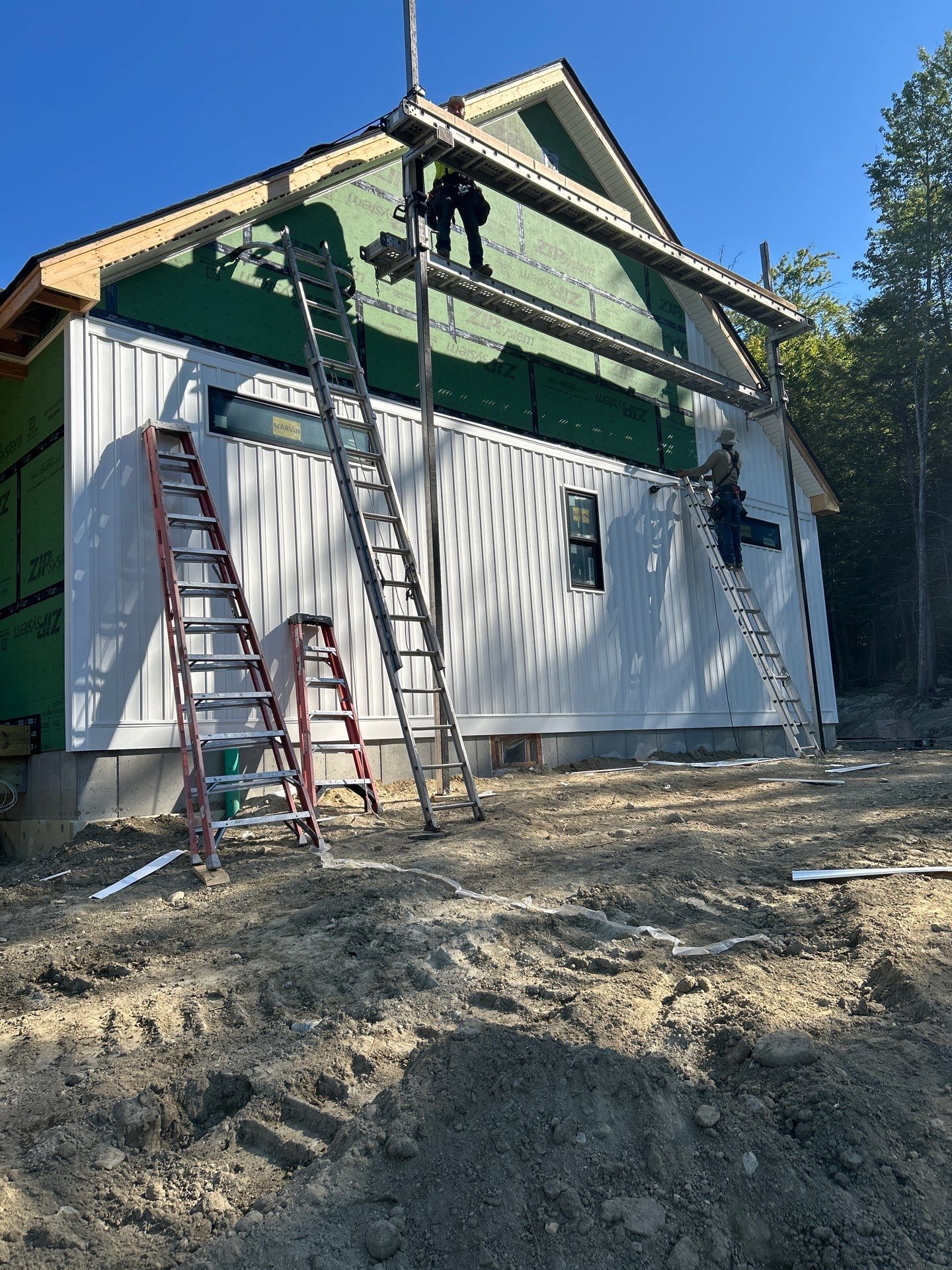 Construction workers installing white siding on house with green insulation visible, multiple ladders and scaffolding equipment on site