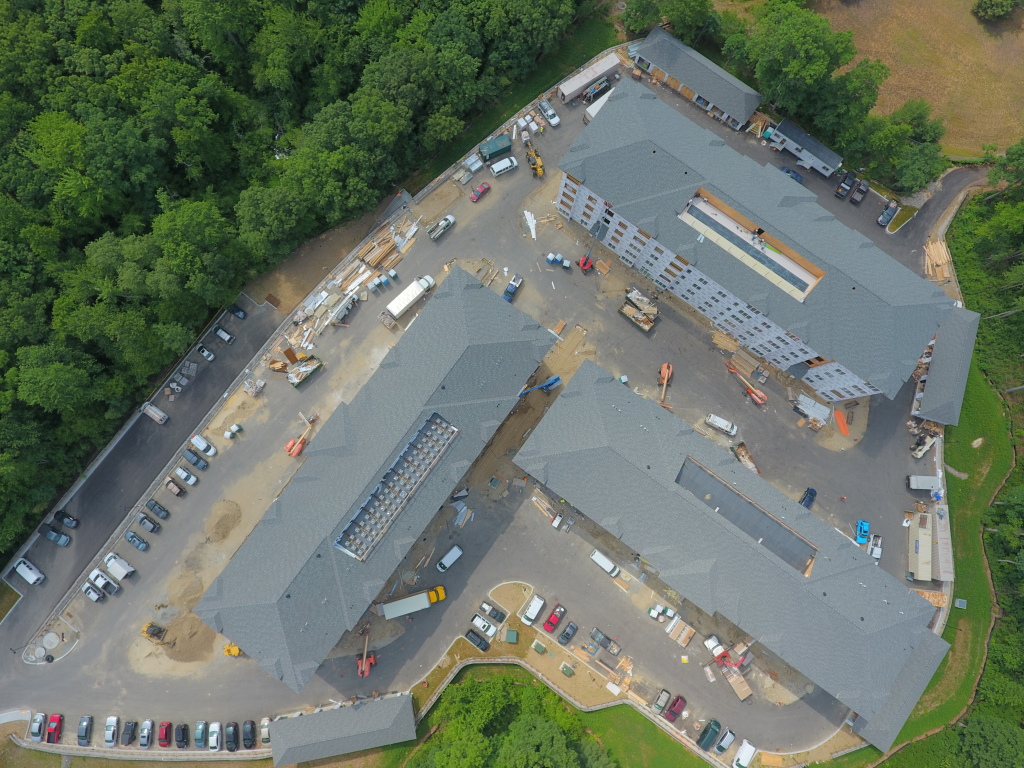 Aerial view of large construction site showing building with gray roof sections, construction vehicles, materials, and worker parking area