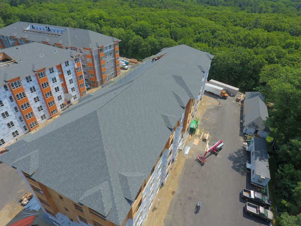 Aerial view of apartment complex construction site with completed buildings, ongoing roofing work, and construction vehicles surrounded by forest