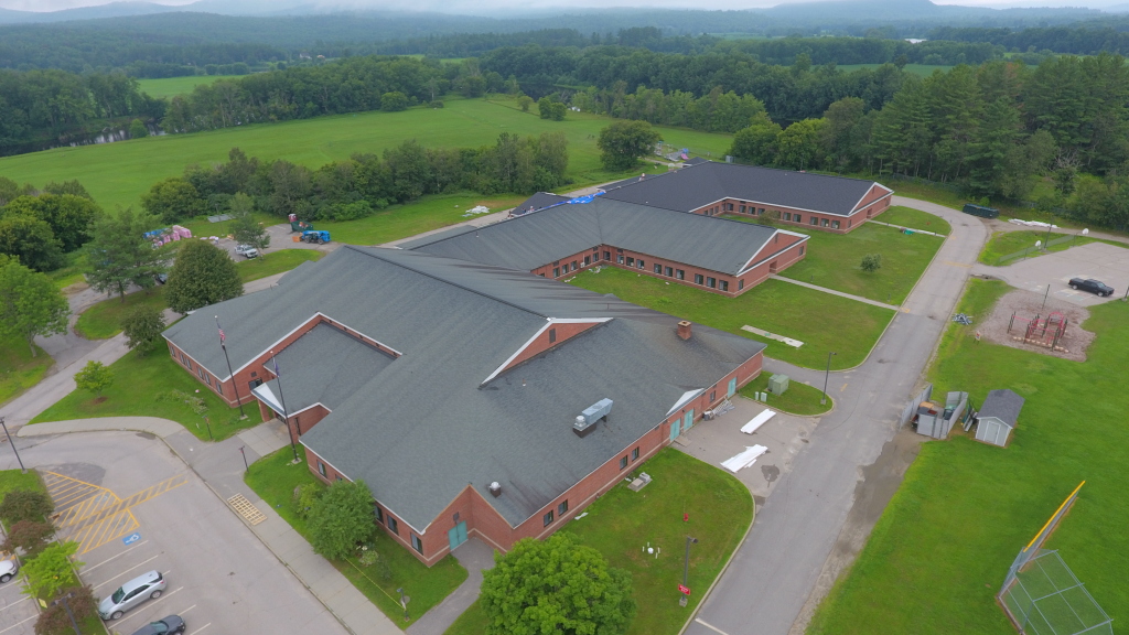 Aerial view of red brick school buildings with dark roofs surrounded by green fields, trees, and mountains in rural setting