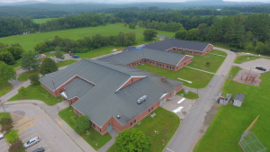 Aerial view of red brick school buildings with dark roofs surrounded by green fields, trees, and mountains in rural setting