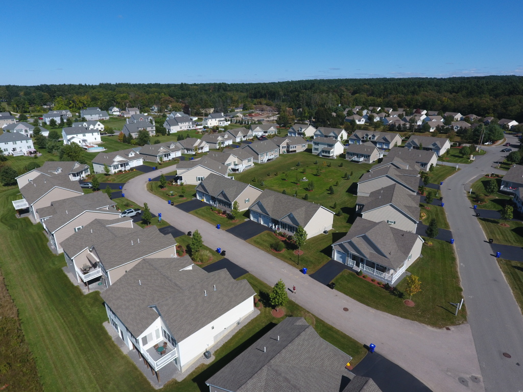 Aerial view of suburban residential neighborhood with modern single-family homes, curved streets, and green lawns surrounded by forests