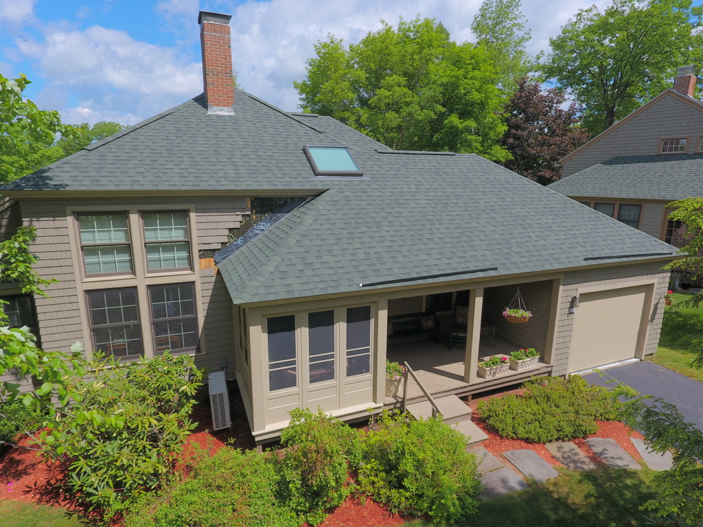 Aerial view of a modern single-story home with gray shingles, covered porch, skylights, and landscaped yard with red mulch