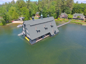 Large Tudor-style boathouse with distinctive twin cupolas built over water, surrounded by lakefront homes and lush green forest