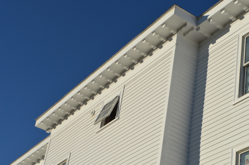 White wooden house exterior with decorative corbels under eaves and open windows against bright blue sky