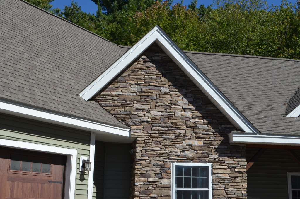 Modern house exterior featuring natural stone gable accent, gray shingle roof, green siding, and wooden garage door surrounded by trees