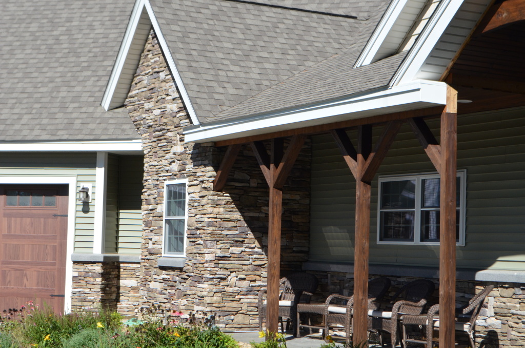 Modern home exterior with stone chimney, covered porch with wooden posts, green siding, and outdoor patio furniture