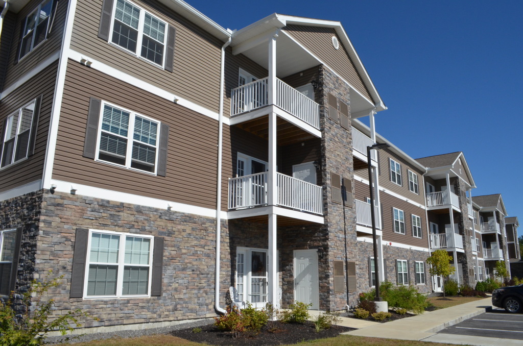 Modern three-story apartment complex with brown siding, stone accents, white balconies, and landscaped front yard under blue sky