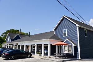 Blue and white commercial building with storefront windows, outdoor seating area with orange umbrella, and parked cars in front
