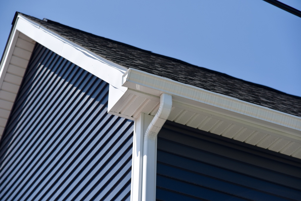 White gutters and downspout on house exterior with dark roof shingles and blue vinyl siding against clear blue sky